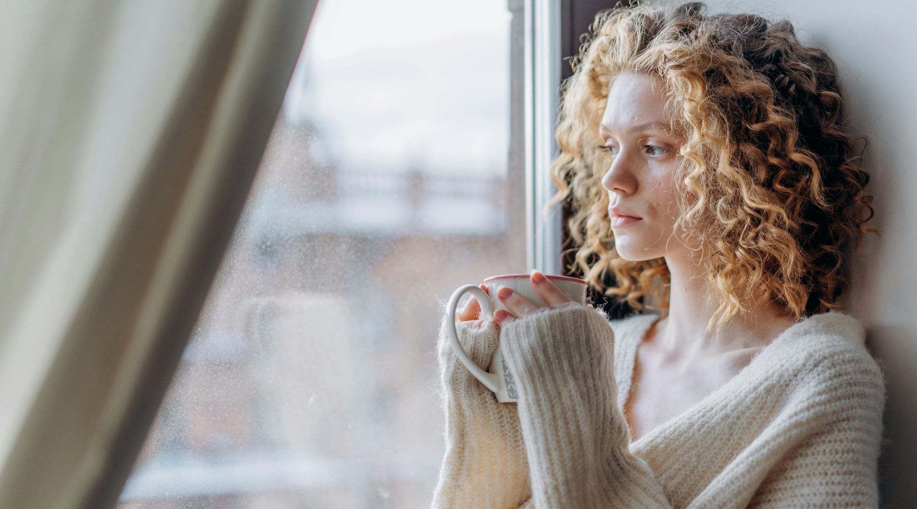 Woman with curly hair sitting at a window holding a cup of coffee, reflecting on her wellness routine and spring reset with CBD products from Thryv Organics.